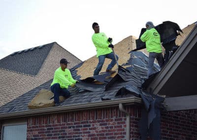 Roofing crew in bright shirts removing shingles from a home's roof on a sunny day.