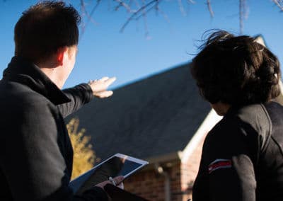 Two individuals discussing a home's roof while one points, holding a tablet. Clear blue sky above.