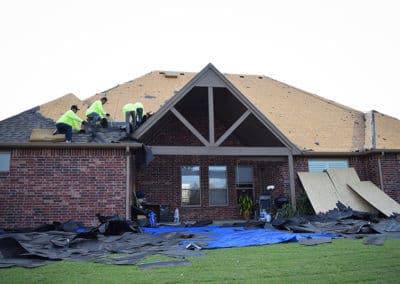 Roofing crew installing new shingles on a house with a brick exterior and yard debris.