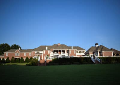Spacious brick mansion with a large lawn, white fences, and clear blue sky.