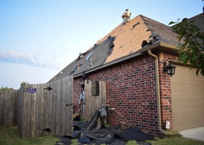 Roof repair in progress on a brick house with a damaged fence and worker on the roof.