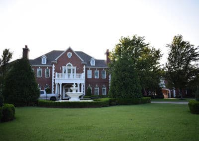 Elegant red brick mansion with a white balcony, surrounded by lush green manicured lawn.