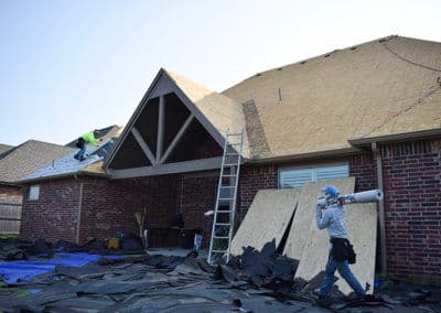 Roofing crew installing new shingles on a house, with a worker carrying supplies below.