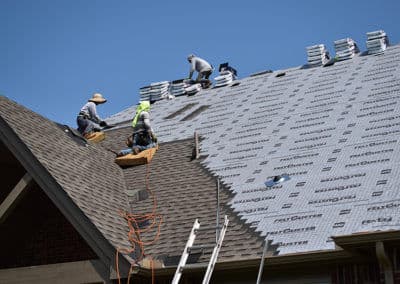 Roofers installing shingles on a house under a clear blue sky.