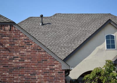 Residential roof with shingles, brick wall, and window under clear blue sky.