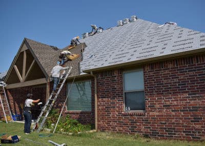Workers repairing a residential roof with ladders and roofing materials under a clear blue sky.