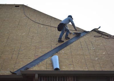 Worker installing roofing material on a sloped roof with a tool in hand.